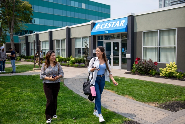 Two students walking away from the Cestar campus doors, talking, causal.