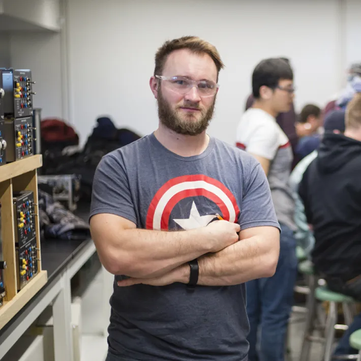 A photo of a student in the electrical lab posing for photo with eletrical equipment and students behind him.