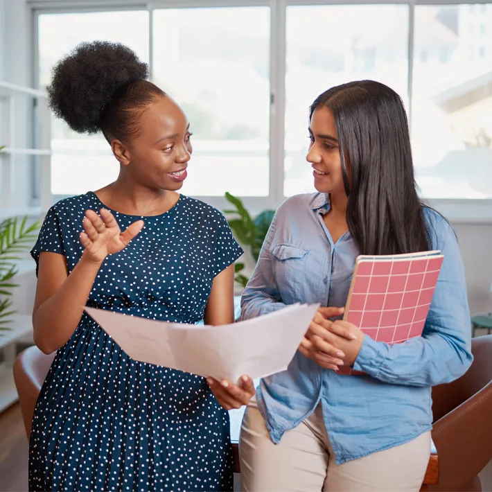 student-mentor-office-stock A photo of an adult mentor talking with a student while holding sheets of paper and notebooks.