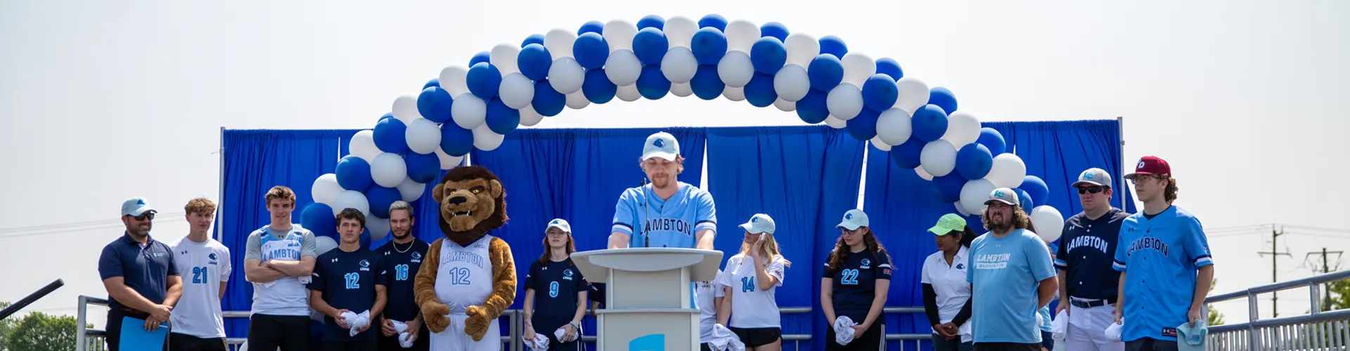 Students and support staff standing on outdoor stage with balloon arch in background.