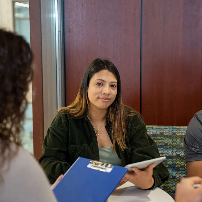 A lifestyle photo of a student sitting at a both holding an ipad with group of people.