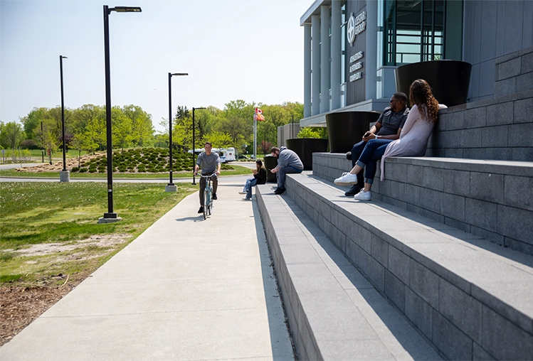 Students lounging outside around the Sarnia Campus entrance, one is riding a bicycle.