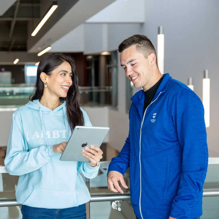 A campus photo of two students standing in a hallway smiling at an ipad.