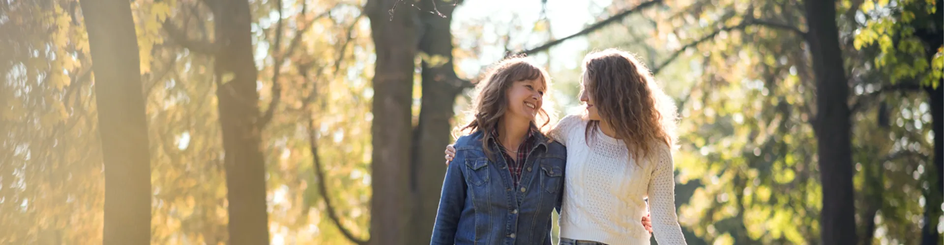 An older and younger woman walking in forested area with arms around each other.