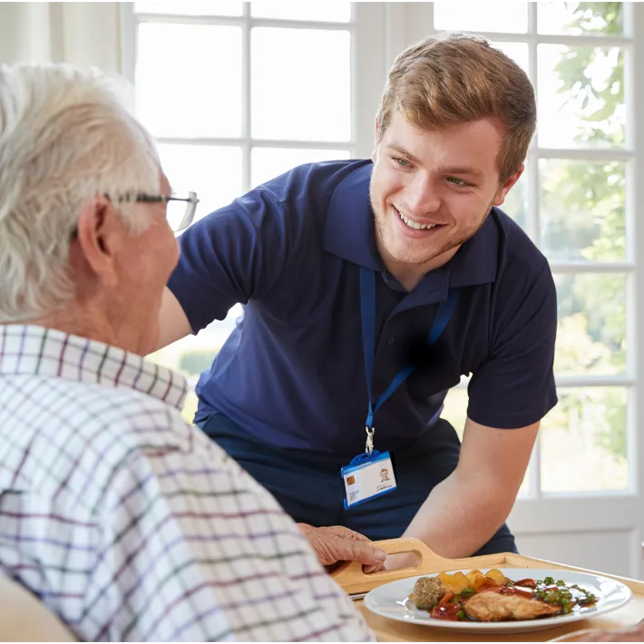 An image of a young resident care member smiling at an older resident with plate of food in front of him.
