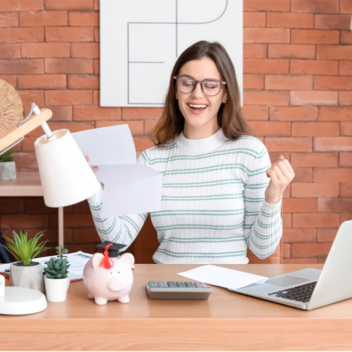 A photo of a young women sitting at desk laughing and smiling with paper in hand and calculator and laptop on desk.