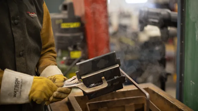 A student picking up welded materials with tongs and wearing safety gloves.