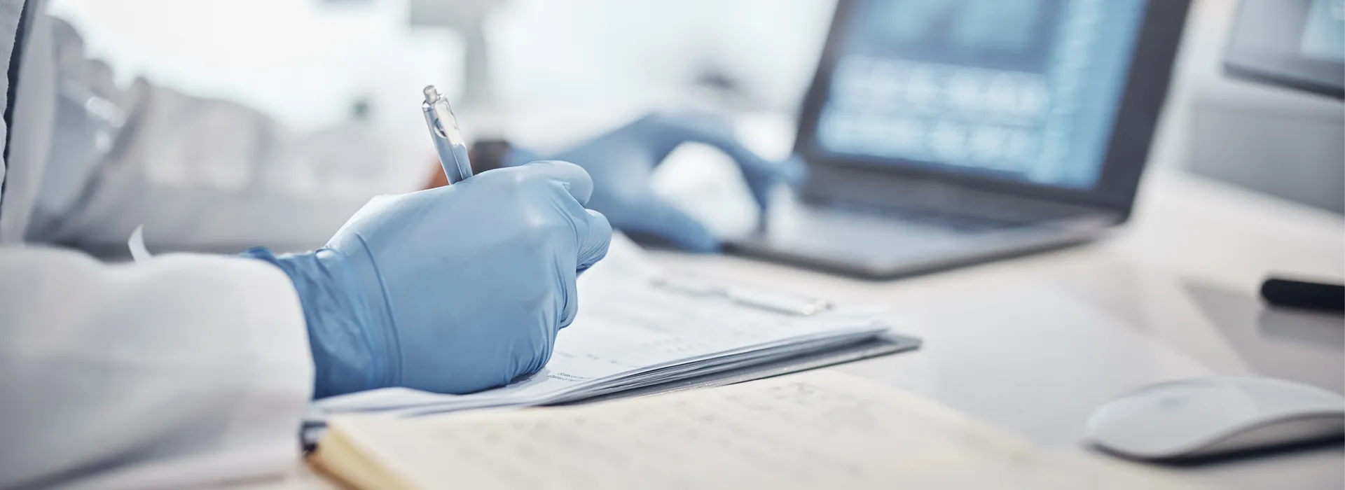 Doctor wearing blue gloves while writing on notepad and on computer.