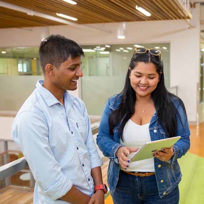 A lifestyle photo of two students standing in hallway looking at ipad on campus and smiling.