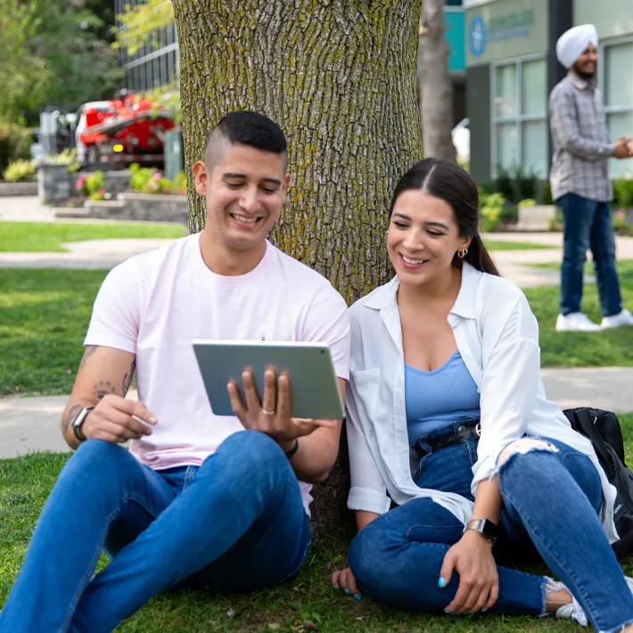 Cestar-Lifestyles-students-international A lifestyle photo of two students wearing white tshirts and blue jeans sitting outside against a tree and looking at ipad together.