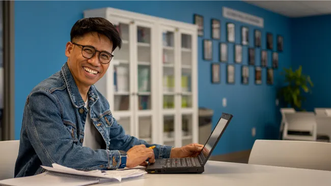 An international student sitting at a desk in a blue room smiling at camera while on computer.