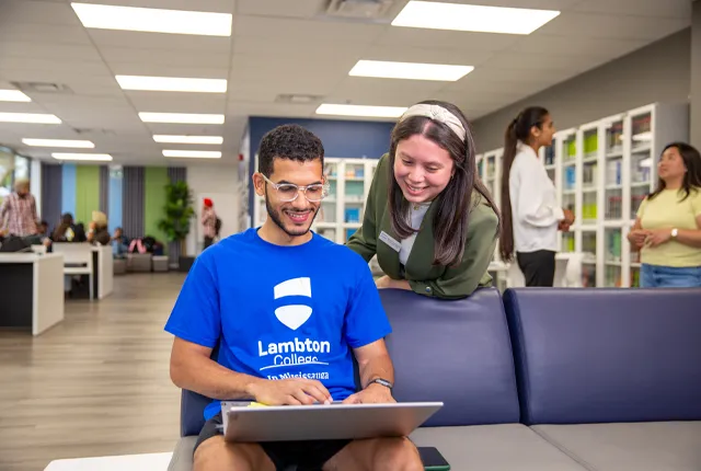 A close up photo of two students in the common room area looking at a computer with other students working and socailizing in the background.