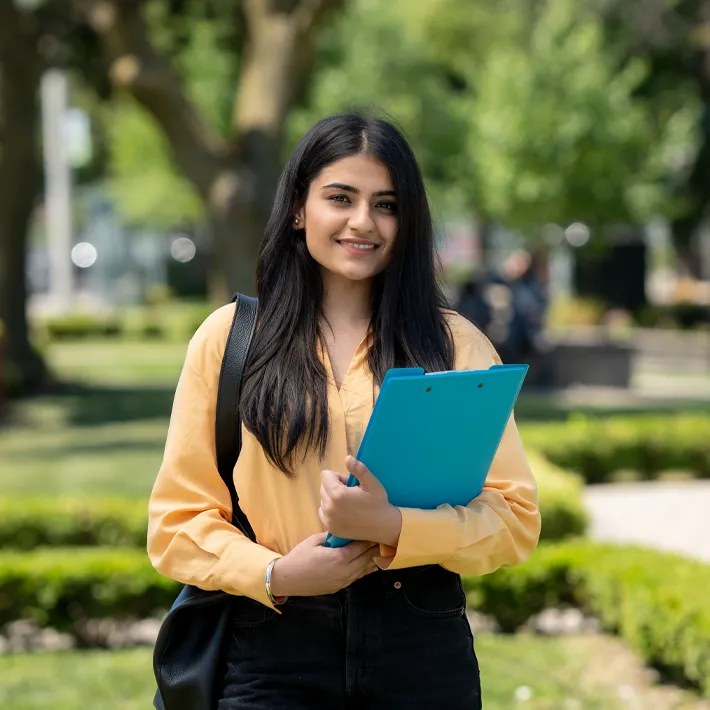 A international student posing for a photo outside on campus grounds holding a binder.