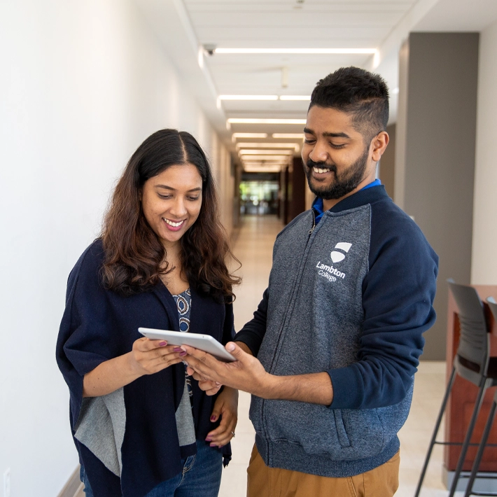 Two international students standing in a hallway on campus looking at an ipad.