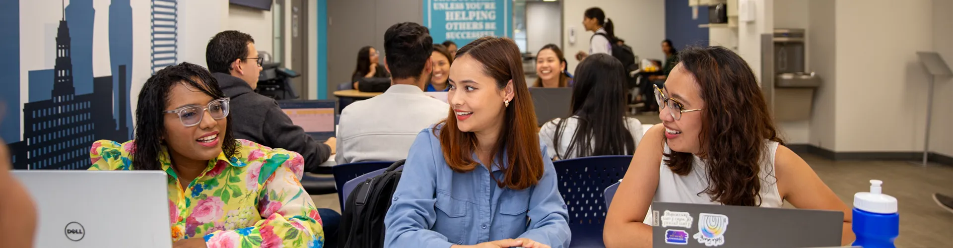 Thre international students working at communal desks with laptops and notes.