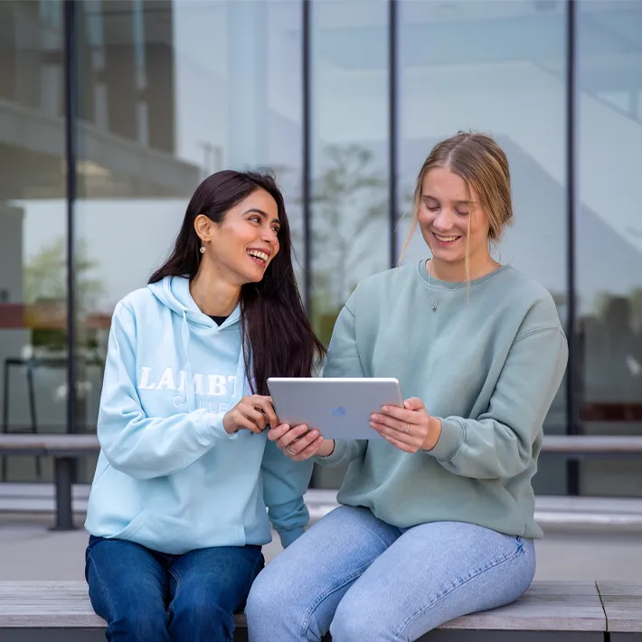 A lifestyle photo of two students, one with dark hair and the other with blonde hair, sitting on a bench outside on campus grounds. They are smiling and holding an iPad.