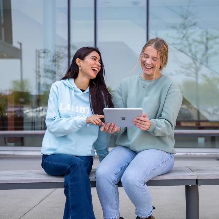 A photo of two international students sitting outside on bench laughing and holding ipad.