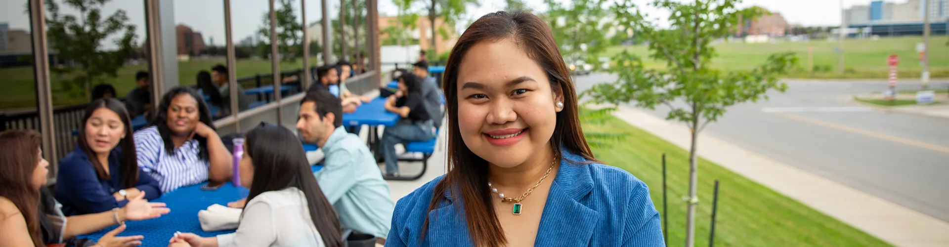 A close up of a student posing oustide for photo while student sitting outside on benches in the background.