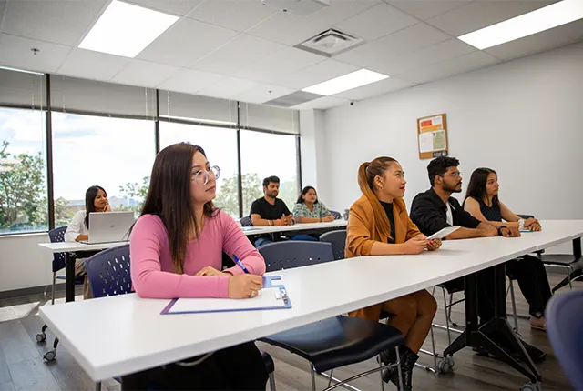 A group of students sitting at desks in a bright classroom with white walls and windows at the back.