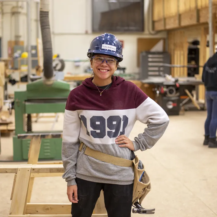 Student in wood shop posing for photo wearing wearing protective gear and tool belt.