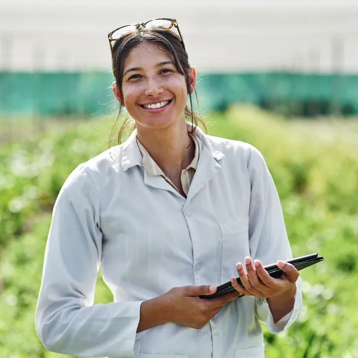 A woman standing in a field of crops holding an ipad and smiling at the camera.