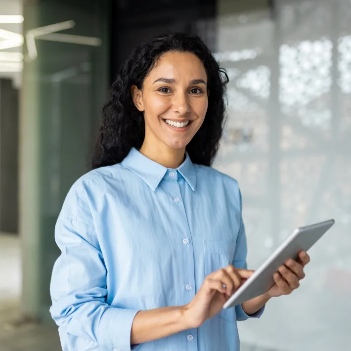 A woman standing in an office wearing a blue shirt and holding an ipad.