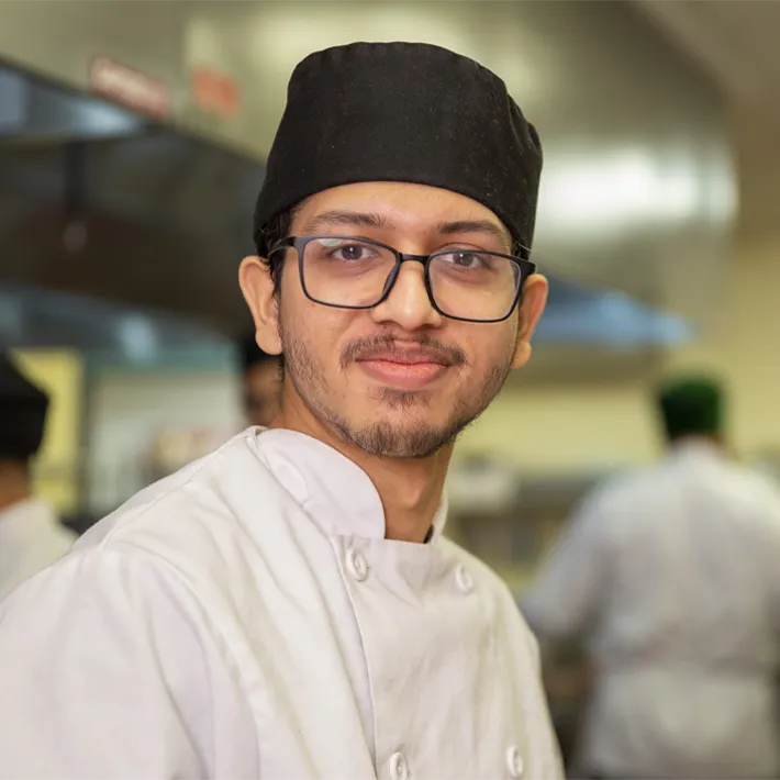 A culinary student posing for photo in kitchen wearing white apron.