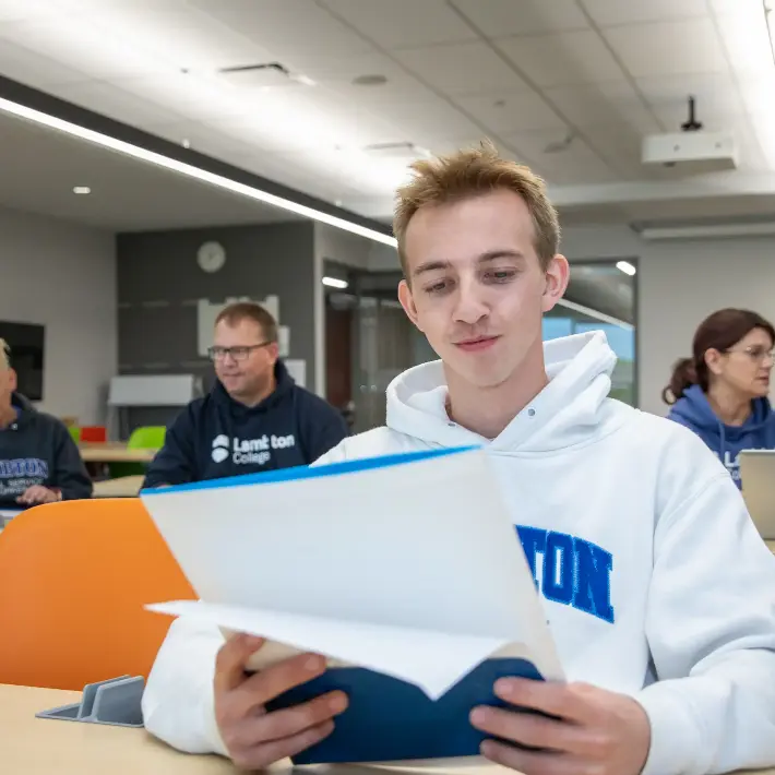 A young student reading his notebook in a classroom, other students in the backyard talking