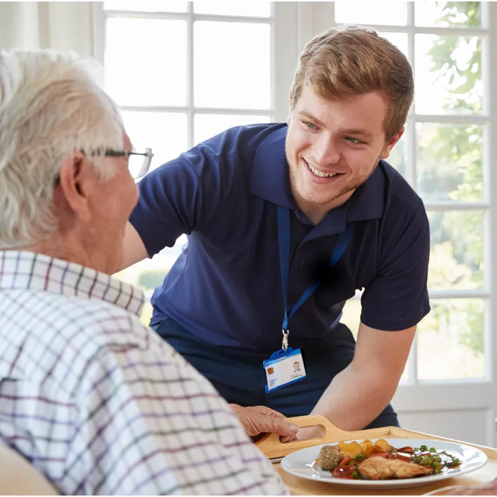 resident-care-training-student-young-old-care-home An image of a young resident care member smiling at an older resident with plate of food in front of him.